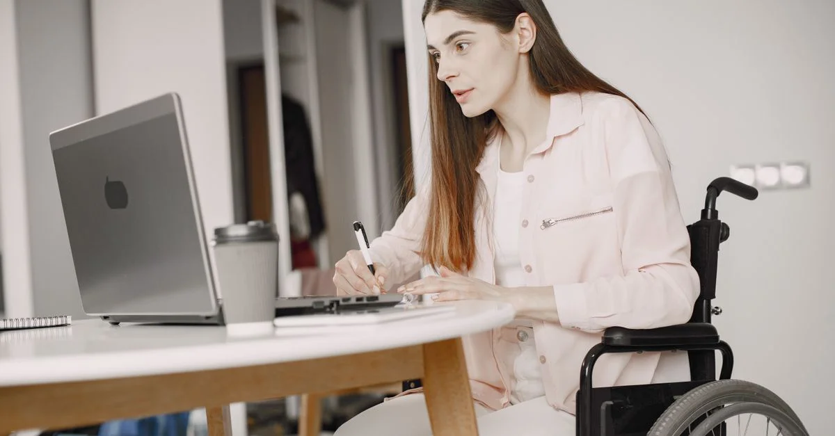 Femme en fauteuil roulant travaillant à distance chez elle avec un ordinateur portable et prenant des notes, mettant en avant le télétravail et l'accessibilité.